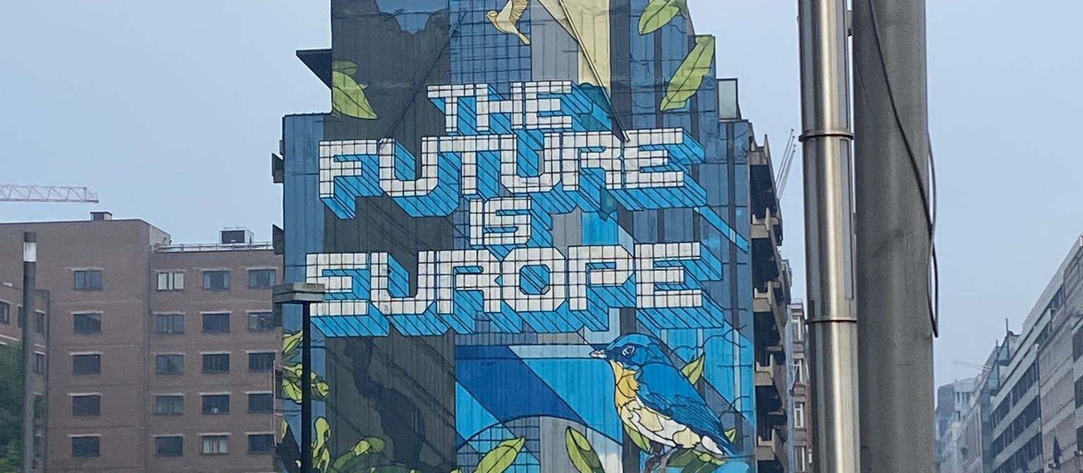 KI generiert: Das Bild zeigt ein großes Wandgemälde an der Fassade eines Gebäudes mit der Aufschrift "THE FUTURE IS EUROPE" und einer Illustration eines Vogels sowie Blättern. Im Hintergrund sind weitere Gebäude und ein Teil einer Straßenlaterne zu sehen.