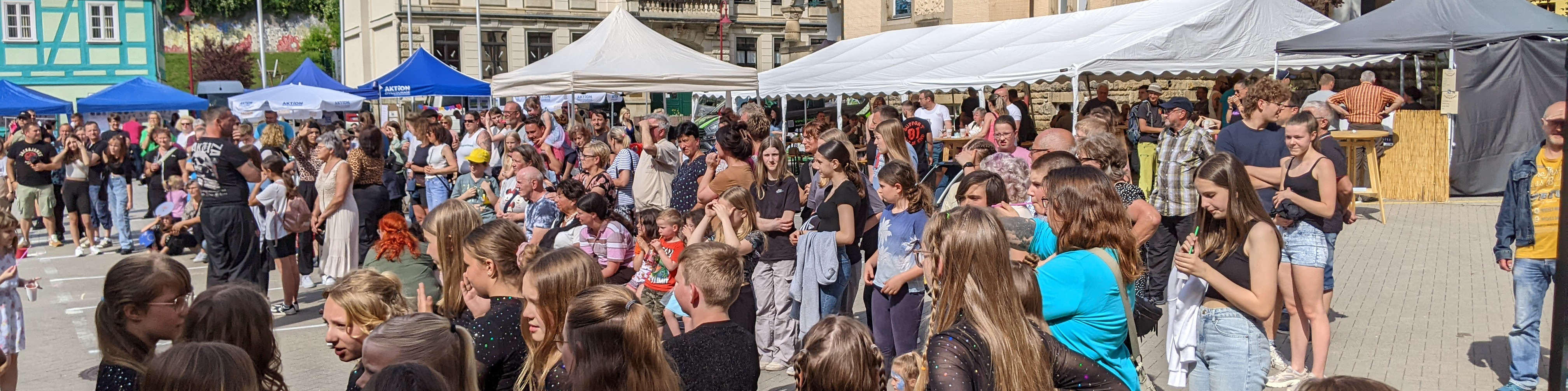 KI generiert: Das Bild zeigt eine belebte Menschenmenge auf einem öffentlichen Platz, möglicherweise während eines Festivals oder einer Veranstaltung, mit mehreren Zelten und Ständen im Hintergrund. Der Hauptinhalt des Bildes ist die Ansammlung von Personen, die sich unterhalten, essen und die Atmosphäre genießen.