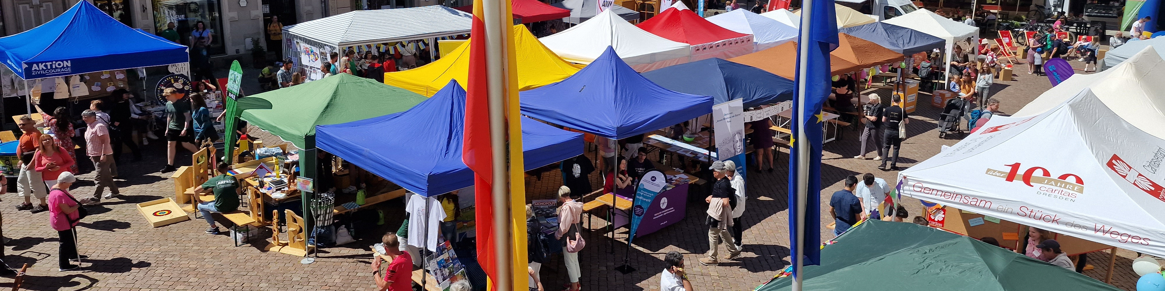 KI generiert: Das Bild zeigt einen belebten Marktplatz mit mehreren farbenfrohen Zelten und Ständen, an denen Menschen verschiedene Waren anbieten und besuchen. Der Hauptinhalt des Bildes ist ein lebhaftes Marktgeschehen mit vielen Besuchern und Ausstellern.