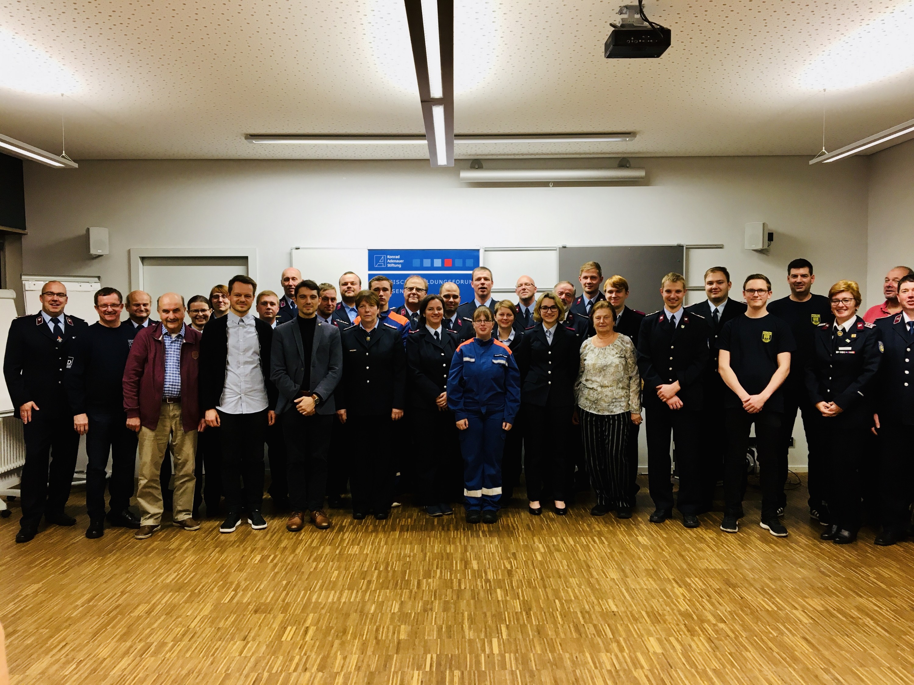 KI generiert: Das Bild zeigt eine Gruppe von Personen, die in einem Raum mit Holzfußboden und weißer Decke stehen. Die meisten tragen Uniformen, was darauf hindeutet, dass es sich um eine offizielle Versammlung oder ein Treffen von Behörden oder Organisationen handeln könnte.