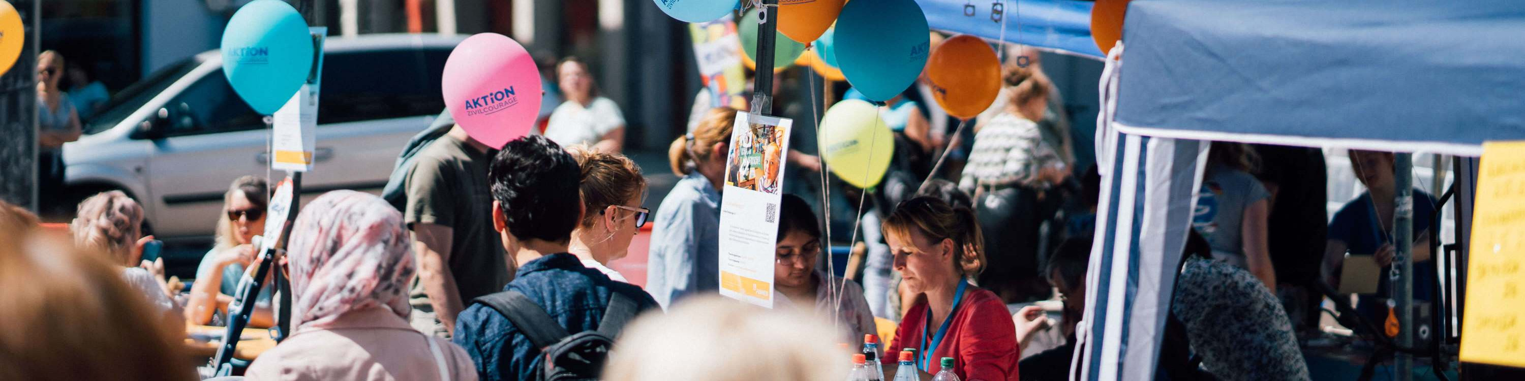 KI generiert: Das Bild zeigt eine belebte Szene mit Menschen, die an einem Stand auf einer Veranstaltung oder einem Markt stehen. Im Vordergrund sind bunte Ballons mit Aufschriften wie "AKTION" zu sehen, die an den Stand gebunden sind.