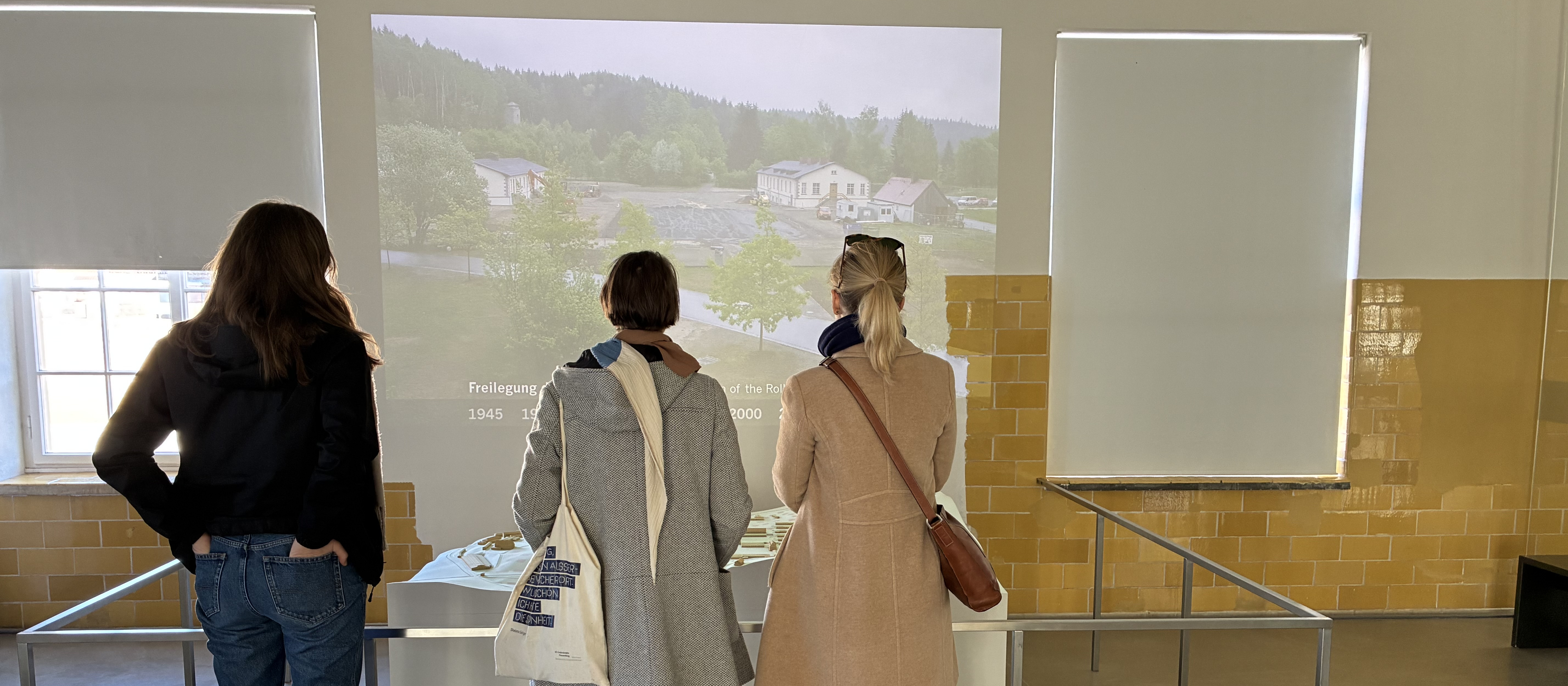 KI generiert: Das Bild zeigt drei Personen, die vor einer Projektion stehen und sich eine Präsentation ansehen, die eine ländliche Landschaft mit Häusern und Bäumen zeigt. Der Hauptinhalt des Bildes ist die Präsentation und die aufmerksamen Zuschauer.