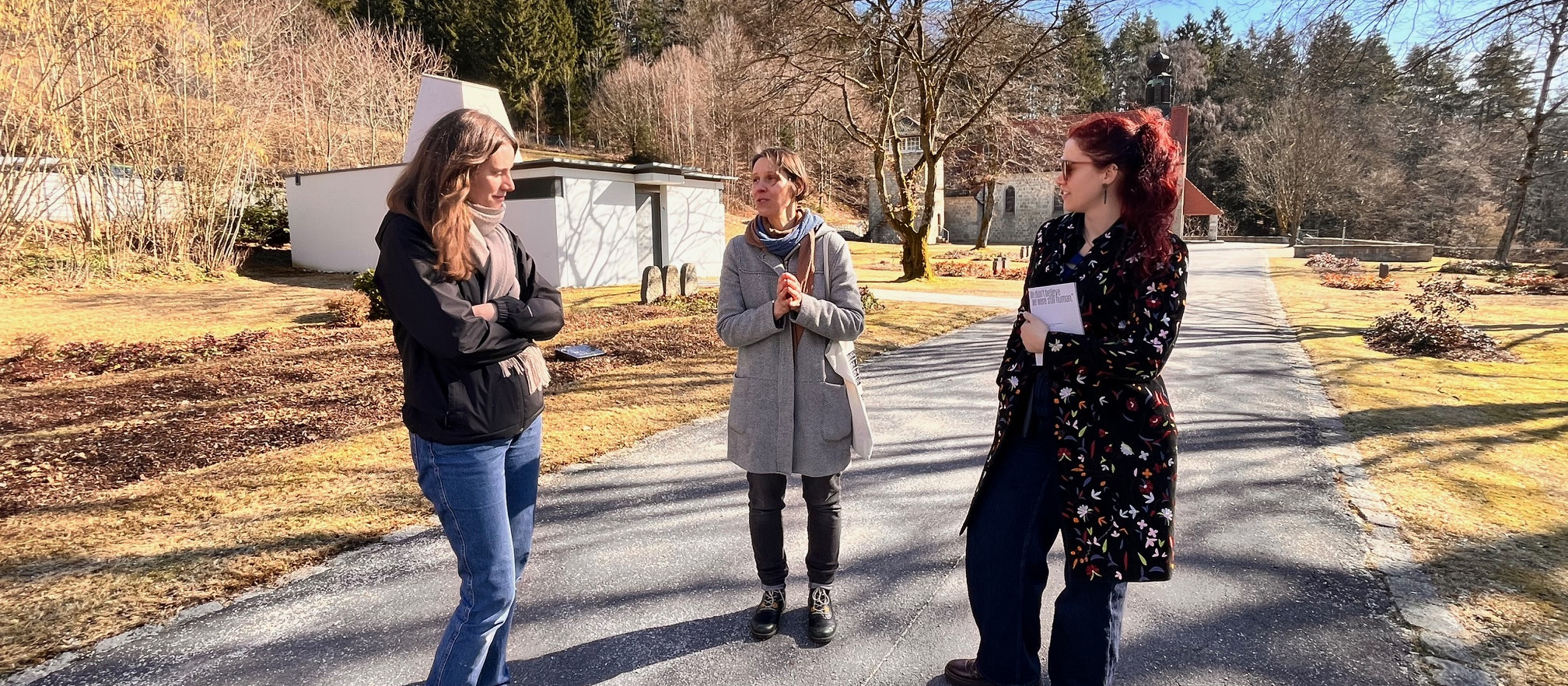 KI generiert: Das Bild zeigt drei Frauen, die auf einem Weg in einer ländlichen, bewaldeten Umgebung stehen und sich unterhalten. Sie halten jeweils ein Blatt Papier in der Hand und scheinen an einer Outdoor-Aktivität oder Führung teilzunehmen.