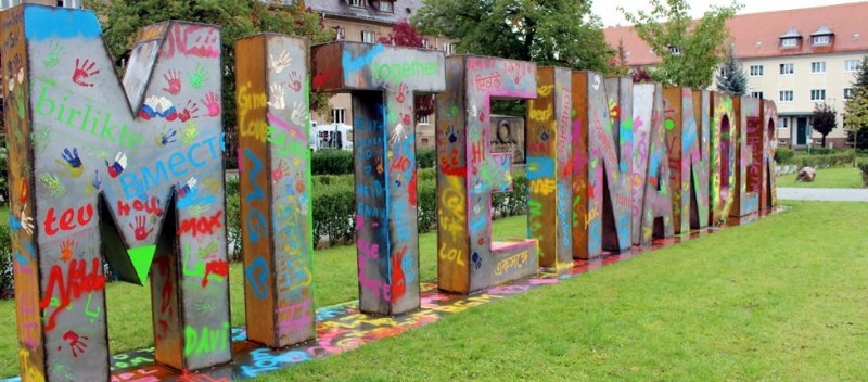 KI generiert: Das Bild zeigt eine bunte, künstlerisch gestaltete Skulptur in Form des Wortes "MITMACHEN" in einem Park. Die Buchstaben sind mit verschiedenen Farben, Handabdrücken und Graffiti verziert.