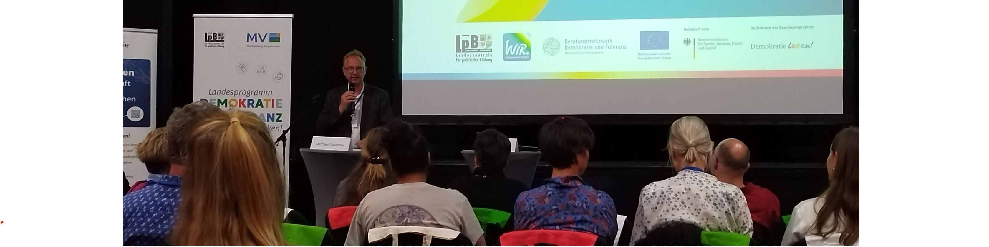KI generiert: Das Bild zeigt eine Präsentation oder einen Vortrag in einem Seminarraum, bei dem ein Mann vor einem Publikum spricht und eine PowerPoint-Präsentation auf einer Leinwand im Hintergrund zu sehen ist. Der Hauptinhalt des Bildes ist die Darstellung einer Bildungs- oder Informationsveranstaltung.