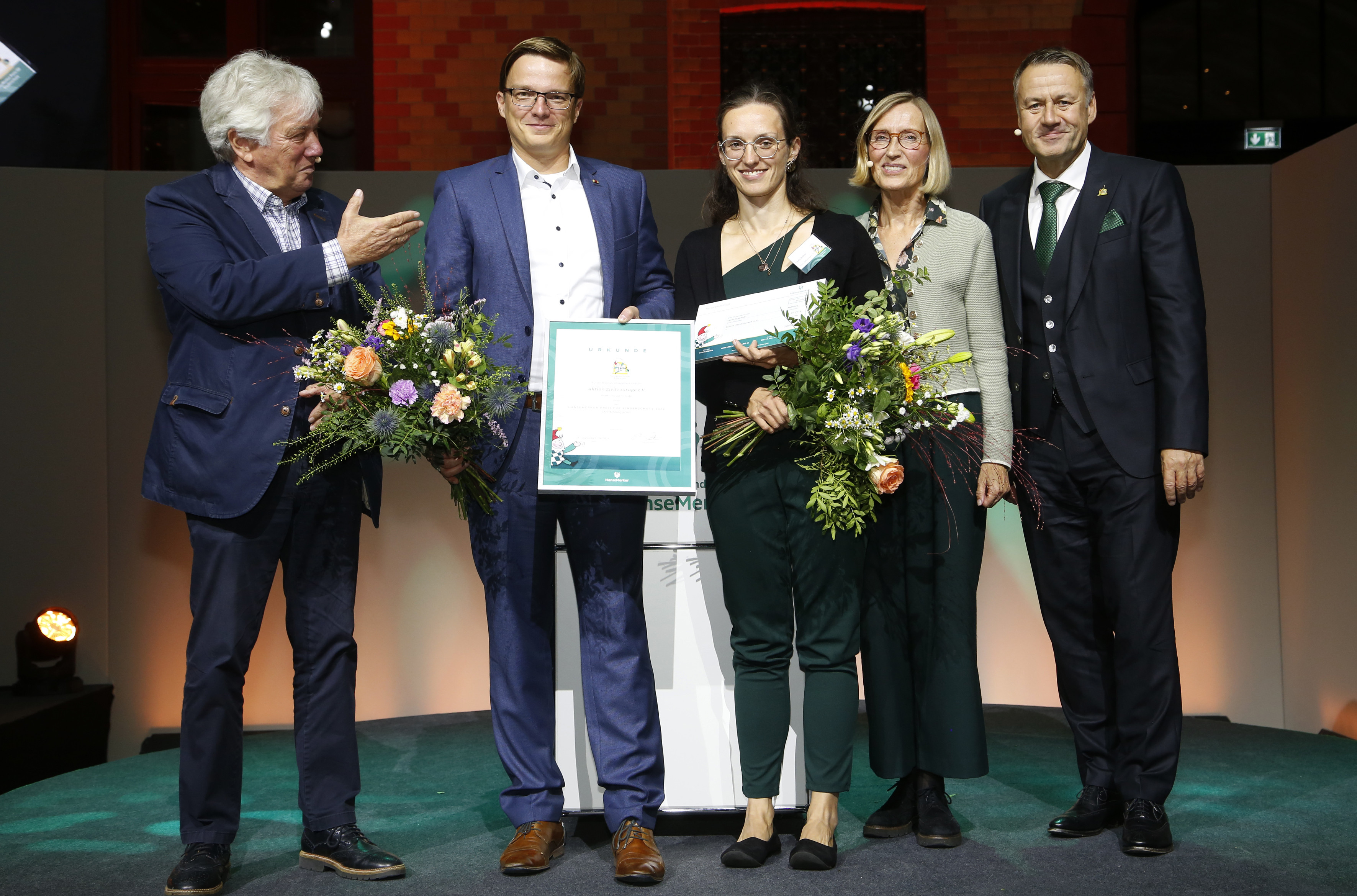 KI generiert: Das Bild zeigt eine Gruppe von fünf Personen auf einer Bühne, die eine Urkunde und Blumensträuße halten, was auf eine Preisverleihung oder Ehrung hindeutet. Die Personen sind feierlich gekleidet und wirken glücklich und stolz.