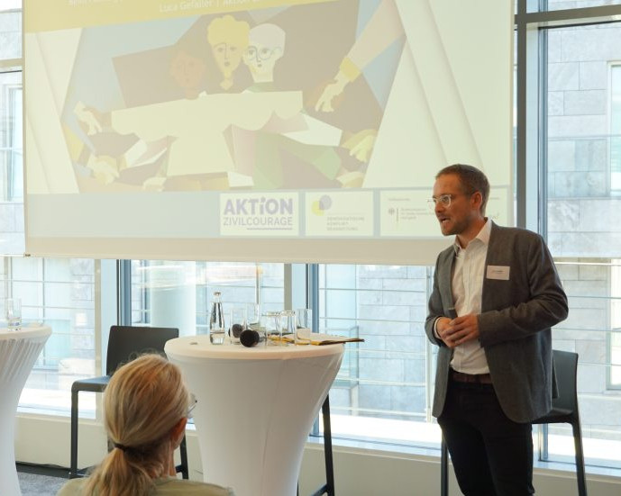 KI generiert: Das Bild zeigt einen Mann, der in einem Konferenzraum vor einem Publikum spricht. Im Hintergrund sind ein Banner mit der Aufschrift "AKTION ZIVILCOURAGE" und ein weiteres Banner mit der Aufschrift "RESPEKT STATT BEDROHUNG!" sichtbar.