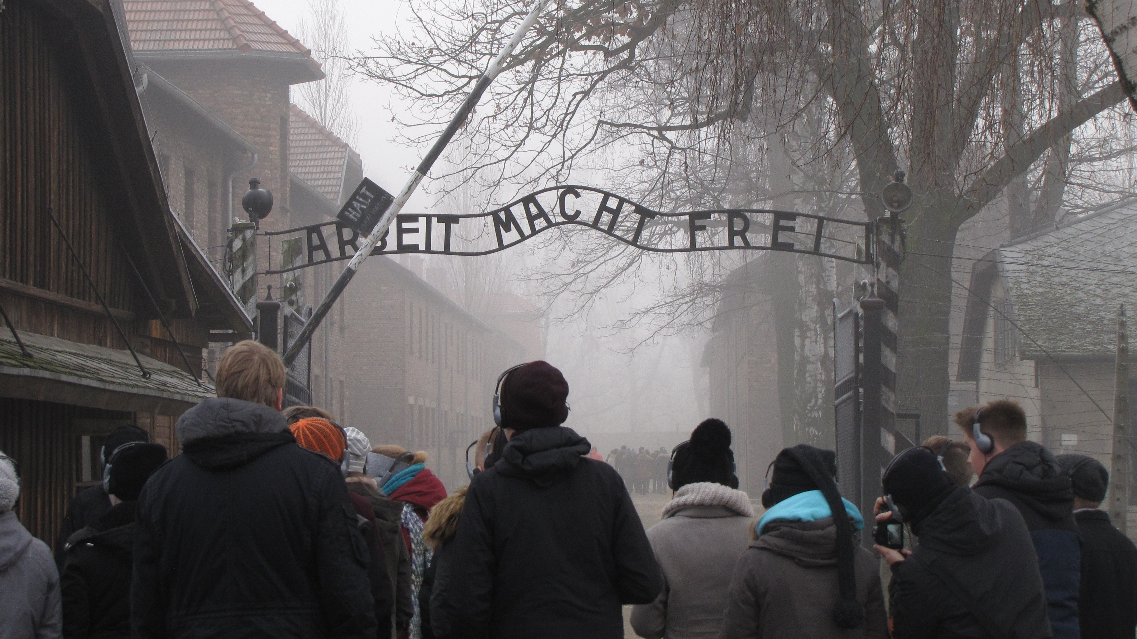 KI generiert: Das Bild zeigt eine Gruppe von Menschen, die unter dem berühmten Tor mit der Aufschrift "Arbeit Macht Frei" (Arbeit macht frei) am Eingang des ehemaligen Konzentrationslagers Auschwitz steht. Der Hauptinhalt des Bildes ist die Gedenkstätte, die an die Opfer des Holocaust erinnert.