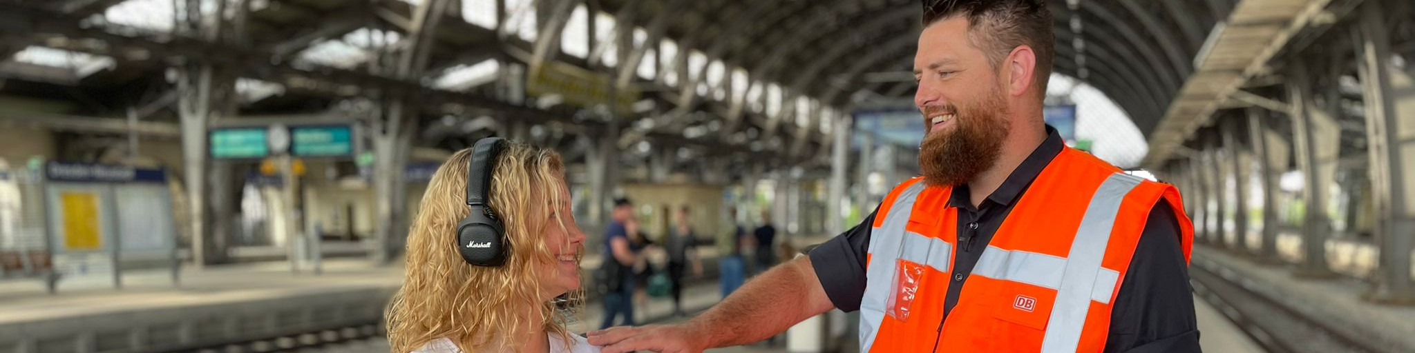 KI generiert: Das Bild zeigt eine Frau mit Kopfhörern und einen Mann in einer orangefarbenen Warnweste, die sich auf einem Bahnsteig unterhalten. Im Hintergrund sind weitere Personen und die Architektur eines Bahnhofs zu erkennen.
