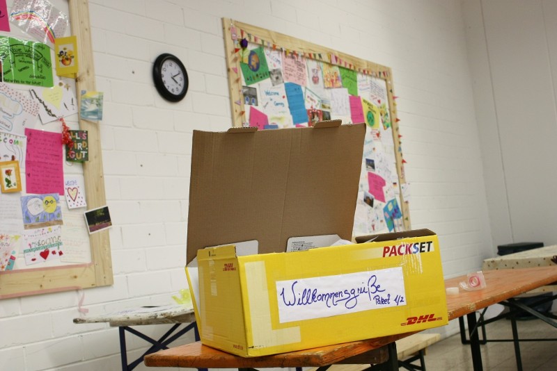 KI generiert: Das Bild zeigt einen Raum mit einem Tisch, auf dem ein offener Karton und ein gelber DHL-Paketkarton mit der Aufschrift "Willkommensgruß" stehen. Im Hintergrund sind eine Pinnwand mit verschiedenen Notizen und Bildern sowie eine Uhr an der Wand zu sehen.