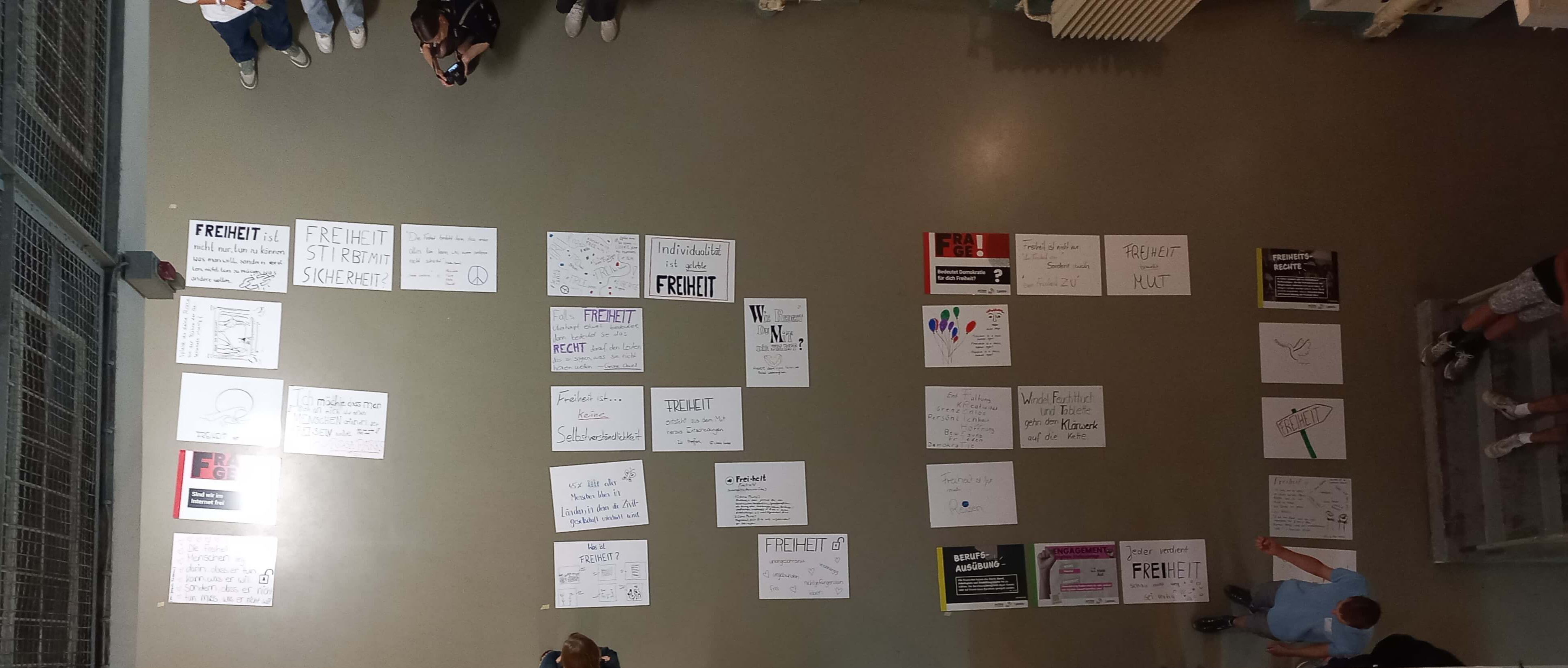 KI generiert: Das Bild zeigt eine Wand, an der zahlreiche handgeschriebene Zettel mit dem Wort "FREIHEIT" (Freiheit) und anderen Botschaften befestigt sind. Im Vordergrund sind Personen, die auf dem Boden sitzen und möglicherweise an einer Veranstaltung oder Diskussion teilnehmen.