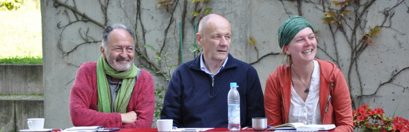 KI generiert: Das Bild zeigt drei Personen, die an einem Tisch sitzen und sich unterhalten. Im Vordergrund sind ein Wasserflasche und einige Tassen zu sehen, während im Hintergrund eine Wand mit Kletterpflanzen und Blumen zu erkennen ist.