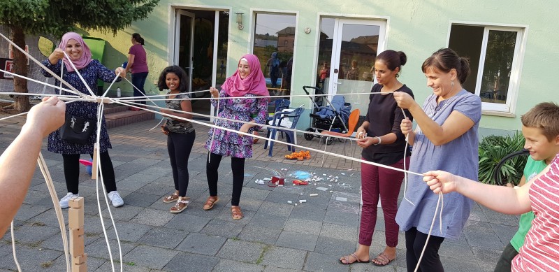 KI generiert: Das Bild zeigt eine Gruppe von Menschen, die gemeinsam an einem Teamspiel teilnehmen, bei dem sie Seile halten, die in der Mitte zusammenlaufen. Der Hauptinhalt des Bildes ist die kooperative Aktivität und das Gemeinschaftsgefühl der Teilnehmer.
