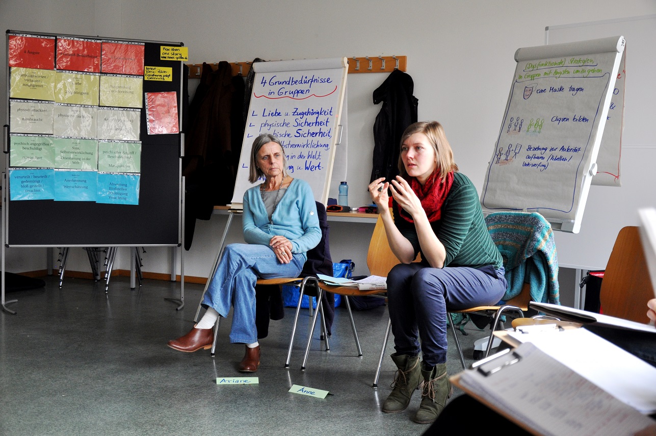 KI generiert: Das Bild zeigt zwei Frauen in einem Seminarraum, die auf Stühlen sitzen und an einer Gruppenarbeit oder Diskussion teilnehmen. Im Hintergrund sind Flipcharts mit Notizen und Plänen zu sehen, die auf eine organisierte Arbeits- oder Lernsituation hinweisen.