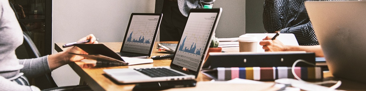 KI generiert: Das Bild zeigt eine Gruppe von Personen, die an einem Tisch sitzen und an Laptops und Tablets arbeiten, wobei Diagramme und Notizen sichtbar sind. Der Hauptinhalt des Bildes ist eine Arbeits- oder Besprechungssituation in einem Büroumfeld.