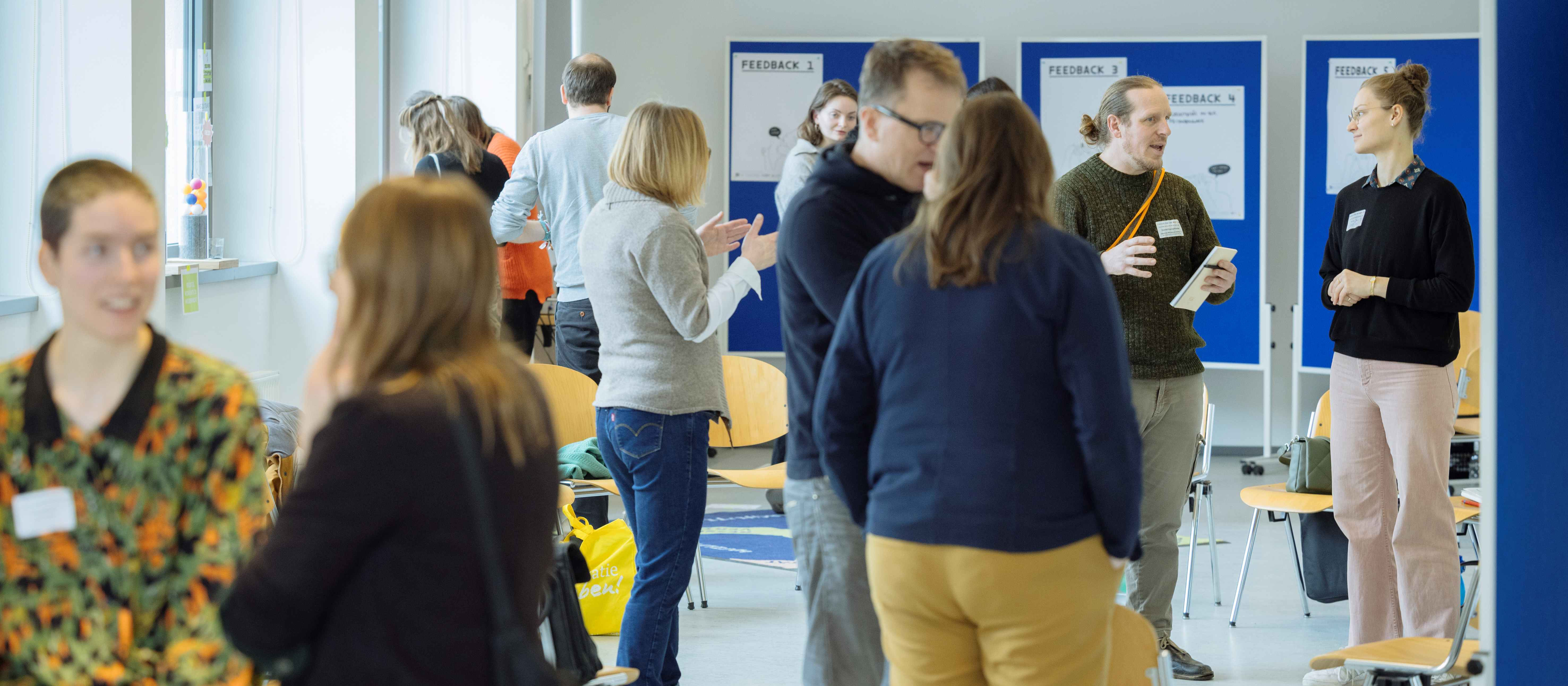 KI generiert: Das Bild zeigt eine Gruppe von Personen in einem hellen Raum, die sich in kleinen Gruppen unterhalten und Notizen machen. Im Hintergrund sind mehrere Flipcharts mit der Überschrift "FEEDBACK" zu sehen, was darauf hindeutet, dass es sich um eine Feedback-Sitzung oder ein Seminar handelt.