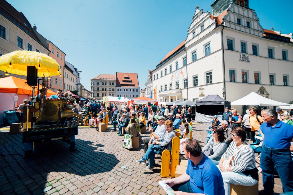 KI generiert: Das Bild zeigt eine belebte Straßenveranstaltung mit vielen Menschen, die auf Bänken sitzen und sich unterhalten, während im Hintergrund historische Gebäude und Marktstände zu sehen sind. Im Vordergrund steht ein altes, gelbes Fahrzeug mit einem großen gelben Schirm.