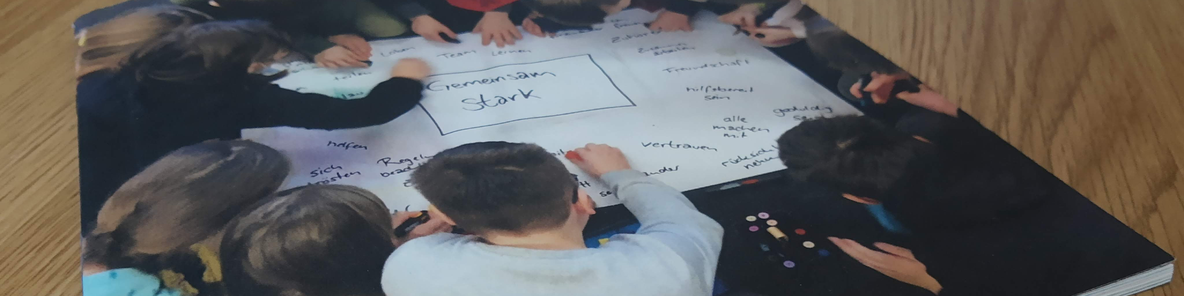 KI generiert: Das Bild zeigt eine Gruppe von Kindern, die gemeinsam an einem großen Blatt Papier arbeiten und verschiedene Begriffe darauf schreiben oder malen. Der Hauptinhalt des Bildes ist eine kreative Gruppenarbeit oder ein Brainstorming, bei dem die Kinder Ideen sammeln und visuell darstellen.