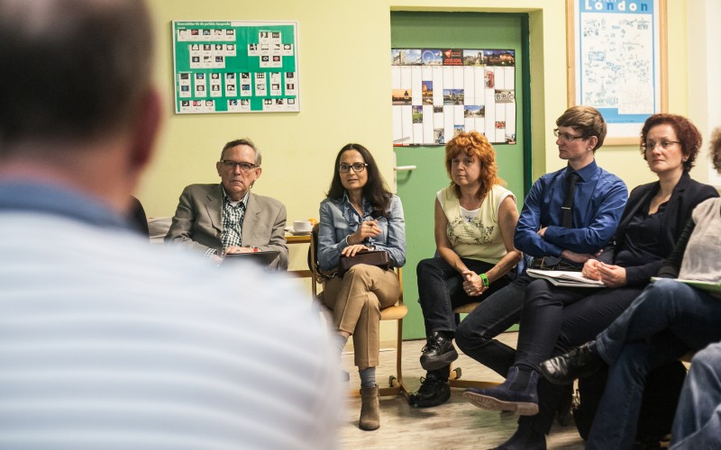KI generiert: Das Bild zeigt eine Gruppe von Personen, die in einem Raum sitzen und einer Diskussion oder einem Vortrag folgen. Der Hauptinhalt des Bildes ist eine kleine Versammlung oder ein Seminar in einem informellen Rahmen.