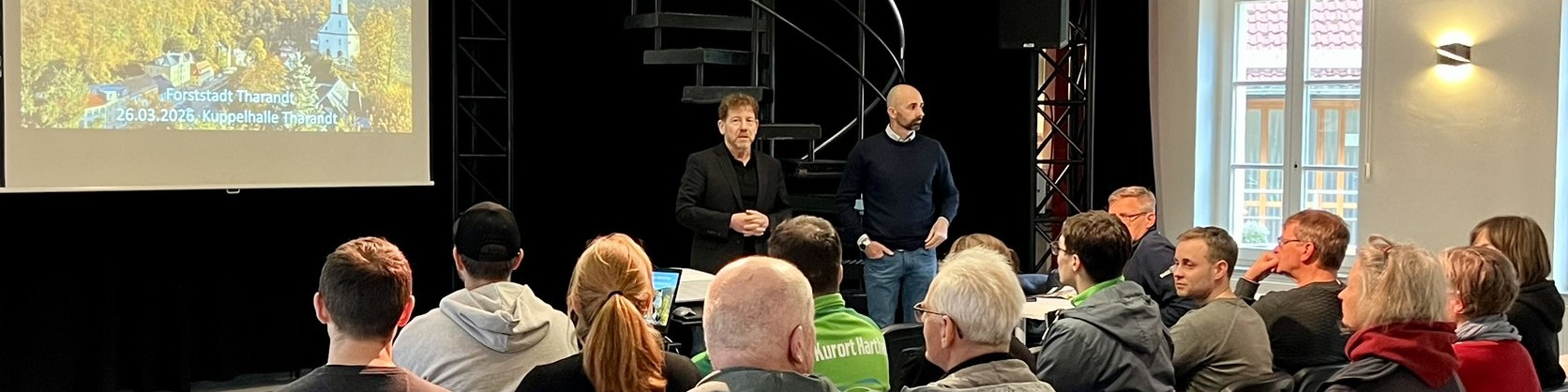 KI generiert: Das Bild zeigt eine Präsentation oder ein Meeting in einem Raum mit mehreren Personen. Zwei Männer stehen vorne und sprechen zu einer Gruppe von Zuhörern, während auf einer Leinwand eine PowerPoint-Präsentation mit dem Titel "Kupferhalte Tausug" und dem Datum "26.03.2025" projiziert wird.