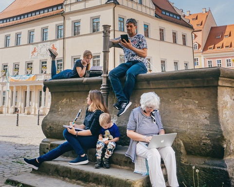 KI generiert: Das Bild zeigt zwei Personen, die auf einem öffentlichen Platz vor historischen Gebäuden sitzen und auf ihre Mobilgeräte schauen. Im Vordergrund ist ein alter Brunnen zu sehen, und im Hintergrund sind traditionelle europäische Häuser mit verzierten Fassaden.