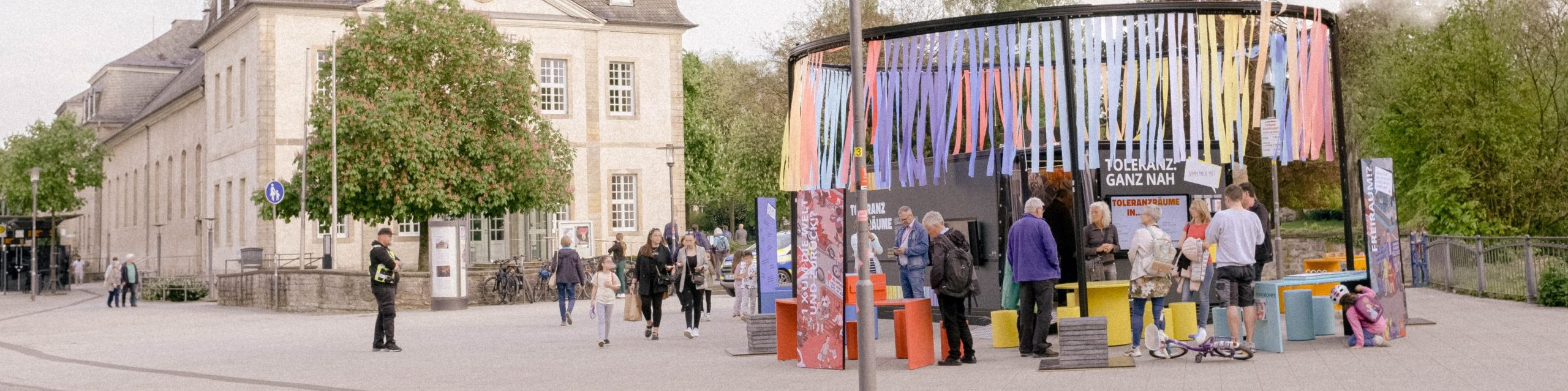 KI generiert: Das Bild zeigt eine belebte Szene mit Menschen, die sich um einen bunten Pavillon mit der Aufschrift "JOCKELS GASTANNAHME GANZ NAH" versammeln, während im Hintergrund ein historisches Gebäude und Bäume zu sehen sind. Der Hauptinhalt des Bildes ist der Pavillon und die Menschen, die sich dort aufhalten.
