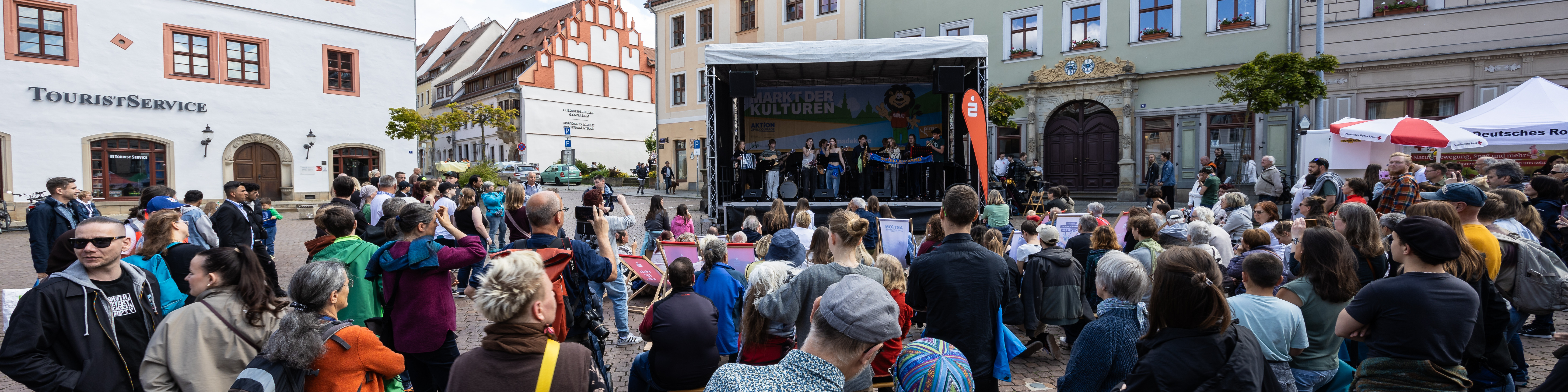 KI generiert: Das Bild zeigt eine belebte Open-Air-Veranstaltung auf einem Platz in einer Stadt mit historischen Gebäuden, bei der eine Band auf einer Bühne auftritt und viele Menschen zuschauen. Der Hauptinhalt des Bildes ist das kulturelle Event mit Live-Musik und einem großen Publikum.
