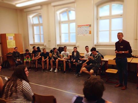 KI generiert: Das Bild zeigt eine Gruppe von Jugendlichen, die in einem Klassenzimmer auf Stühlen sitzen und einem Mann zuhören, der vor ihnen steht und spricht. Der Hauptinhalt des Bildes ist eine Unterrichts- oder Diskussionssituation in einer schulischen Umgebung.