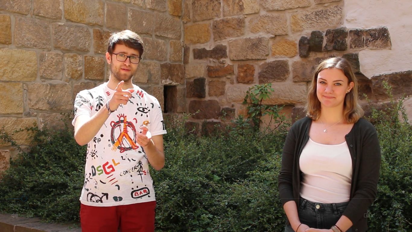 KI generiert: Das Bild zeigt zwei Personen, einen Mann und eine Frau, die vor einer alten Steinmauer und einem Busch stehen. Der Mann trägt ein bunt bedrucktes T-Shirt und hält einen Gegenstand in der Hand, während die Frau ein weißes Oberteil und eine dunkle Jacke trägt.