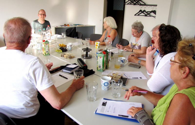 KI generiert: Das Bild zeigt eine Gruppe von Personen, die um einen großen Tisch sitzen und sich in einer Besprechung oder einem Workshop befinden. Auf dem Tisch stehen verschiedene Getränke, Snacks und Arbeitsmaterialien wie Notizblöcke und Stifte.