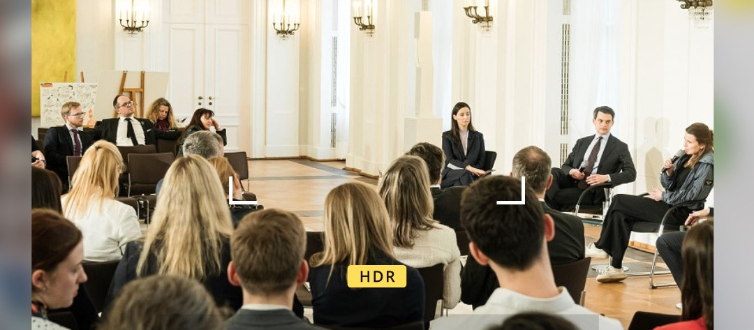 KI generiert: Das Bild zeigt eine formelle Diskussion oder ein Panel in einem hellen, eleganten Raum mit mehreren Teilnehmern, die auf der Bühne sitzen und einem Publikum, das ihnen zuhört. Der Hauptinhalt des Bildes ist eine organisierte Veranstaltung, möglicherweise eine Konferenz oder ein Seminar.