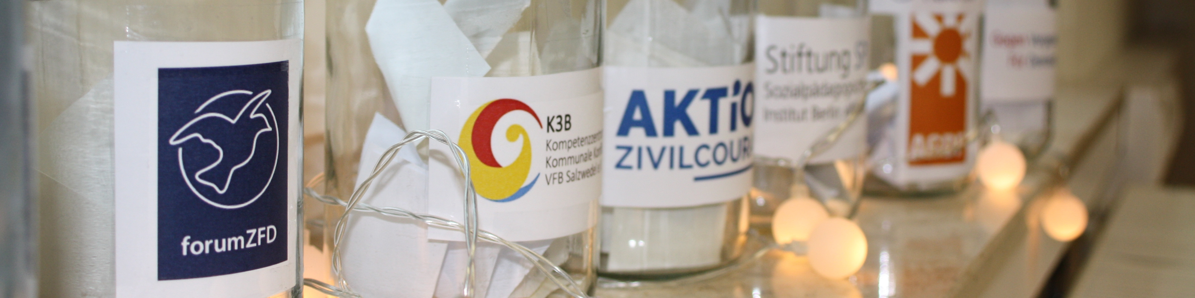 KI generiert: Das Bild zeigt mehrere Gläser mit Aufklebern verschiedener Organisationen und Logos, darunter "forumZFD", "K3B" und "AKTION ZIVILCOURAGE". Im Hintergrund sind warme Lichter zu sehen, die eine gemütliche Atmosphäre erzeugen.