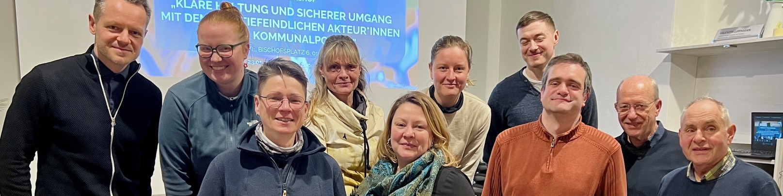 KI generiert: Das Bild zeigt eine Gruppe von zwölf Personen, die gemeinsam in einem Raum posieren. Im Hintergrund ist ein Banner mit Text zu sehen, der auf eine Veranstaltung oder ein Treffen hinweist.