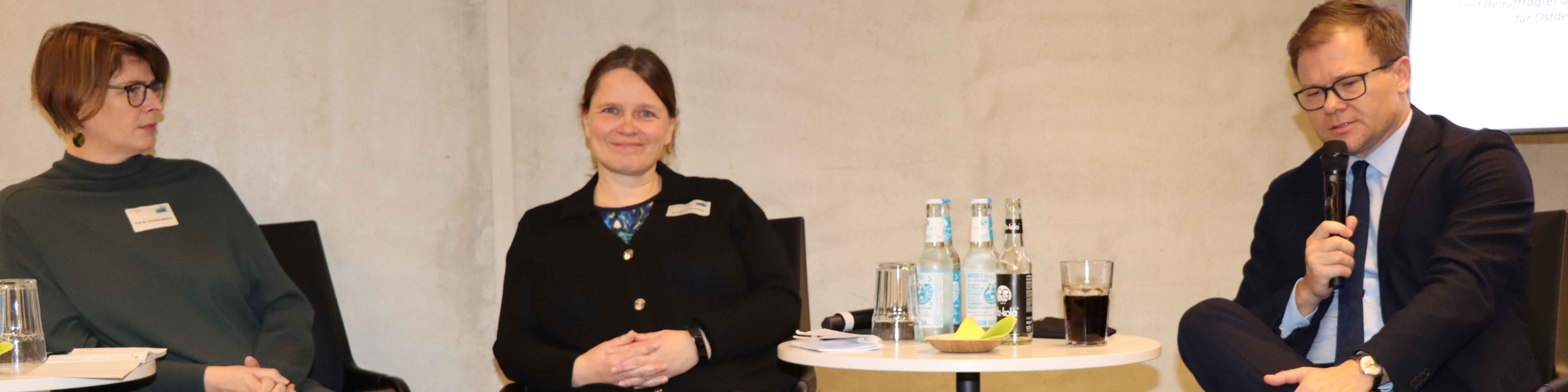 KI generiert: Das Bild zeigt drei Personen, die an einem Tisch sitzen und sich in einer Diskussion oder einem Panel befinden. Auf dem Tisch stehen Wasserflaschen, Gläser und ein Mikrofon, was auf eine formelle Veranstaltung oder Konferenz hindeutet.