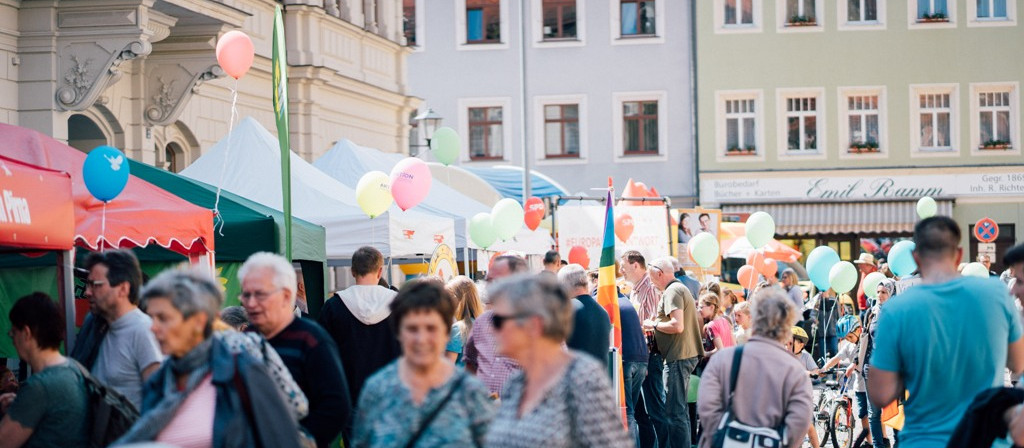 KI generiert: Das Bild zeigt eine belebte Straßenveranstaltung oder ein Fest mit vielen Menschen, bunten Ballons und verschiedenen Ständen. Im Hintergrund sind historische Gebäude zu sehen, die auf einen europäischen Ort hindeuten.