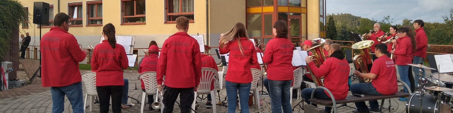 KI generiert: Das Bild zeigt eine Gruppe von Musikern in roten Shirts, die im Freien ein Konzert geben. Die Musiker spielen verschiedene Instrumente, darunter Trompeten, Klarinetten und Schlagzeug.
