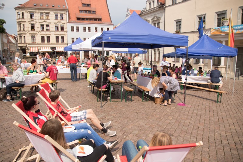 KI generiert: Das Bild zeigt eine belebte Außenveranstaltung auf einem gepflasterten Platz mit mehreren Menschen, die an Tischen unter blauen Zelten sitzen und sich unterhalten. Im Vordergrund entspannen einige Personen auf Liegestühlen.