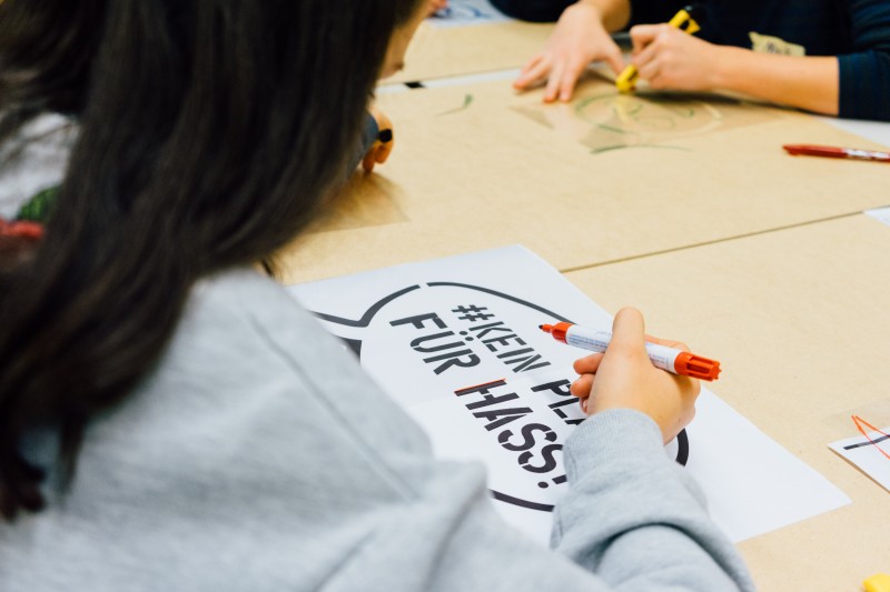 KI generiert: Das Bild zeigt eine Person, die mit einem roten Marker auf einem Blatt Papier schreibt, auf dem bereits der Text "#KEIN PLATZ FÜR HASS!" zu sehen ist. Im Hintergrund sind weitere Personen, die ebenfalls an einem Tisch sitzen und mit verschiedenen Materialien arbeiten.