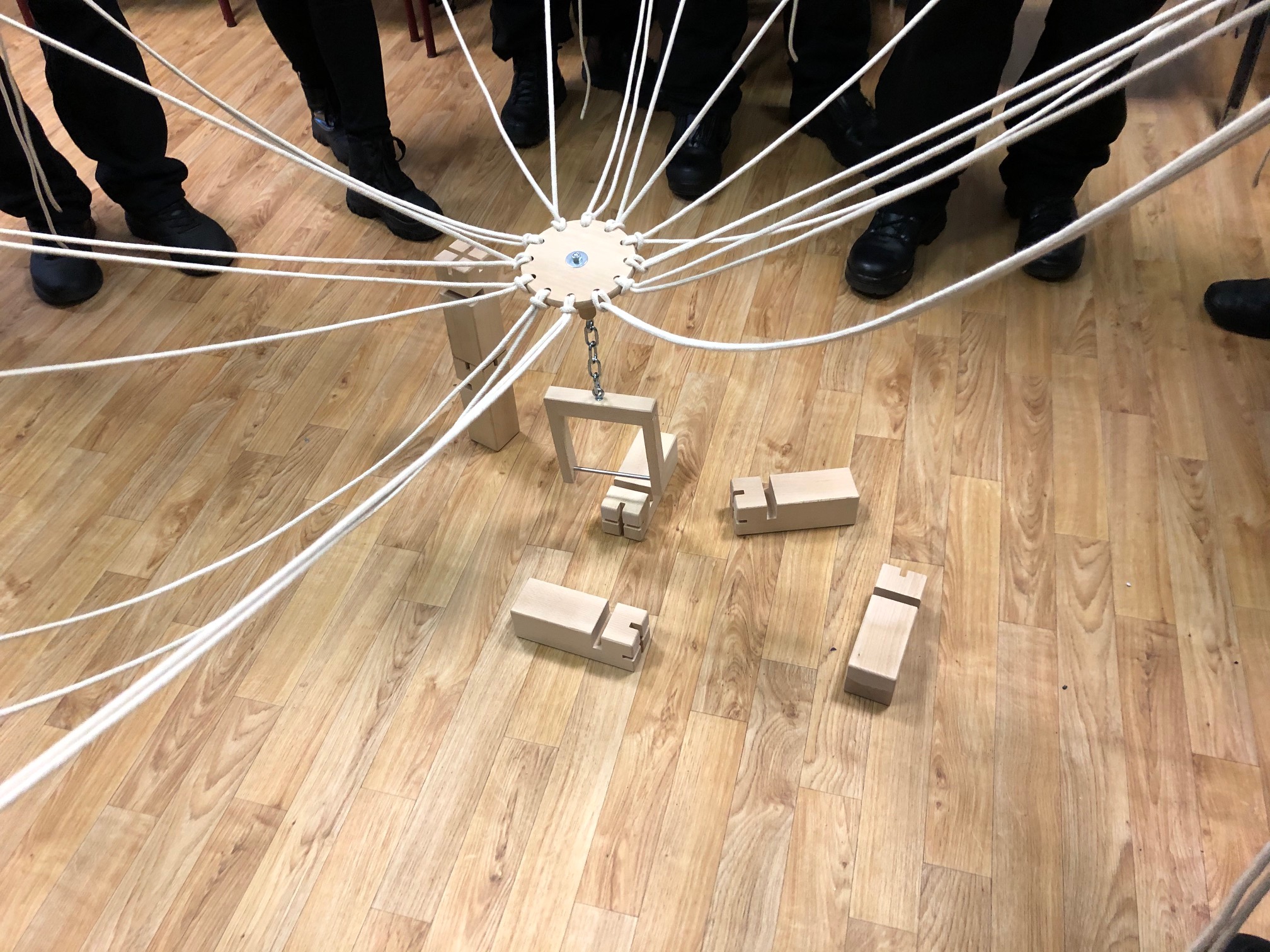 KI generiert: Das Bild zeigt eine Gruppe von Personen, die um eine selbstgebaute Konstruktion aus Holz und Schnüren stehen, die wie ein mechanisches Modell oder eine Kunstinstallation aussieht. Der Hauptinhalt des Bildes ist diese Konstruktion, die aus einem zentralen Rad mit vielen Schnüren besteht, die zu verschiedenen Holzteilen auf dem Boden führen.