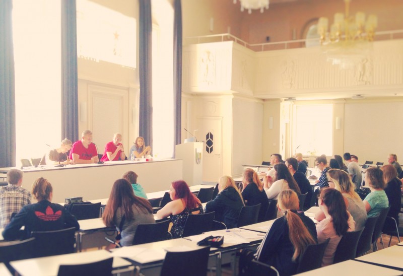 KI generiert: Das Bild zeigt einen gut beleuchteten, großen Raum, der wie ein Hörsaal oder ein Sitzungssaal aussieht, mit vielen Personen, die an langen Tischen sitzen und einer Gruppe von Personen auf einem Podium zuhören. Der Hauptinhalt des Bildes ist eine Versammlung oder ein Seminar, bei dem eine Diskussion oder Präsentation stattfindet.