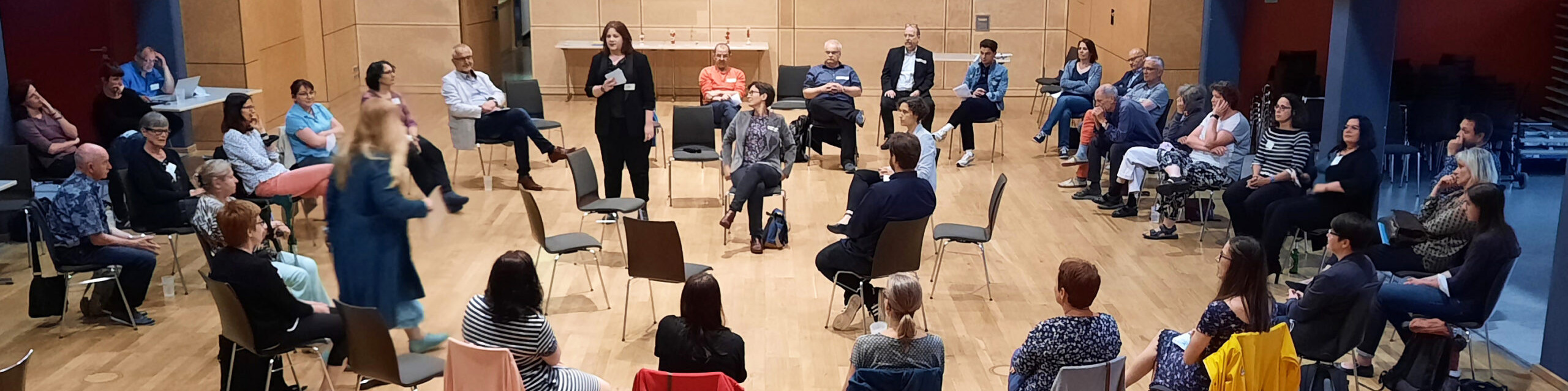 KI generiert: Das Bild zeigt eine Gruppe von Menschen, die in einem Kreis in einem Raum mit Holzvertäfelung sitzen und sich anscheinend in einer Diskussion oder einem Workshop befinden. Der Hauptinhalt des Bildes ist eine interaktive Gruppenaktivität oder ein Meeting.
