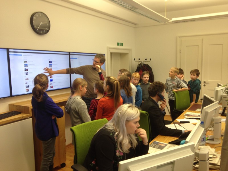 KI generiert: Das Bild zeigt eine Gruppe von Kindern und Erwachsenen in einem Büro oder Klassenzimmer, wobei ein Mann vor einem großen Bildschirm steht und etwas erklärt. Der Hauptinhalt des Bildes ist eine interaktive Lernsituation oder Präsentation.