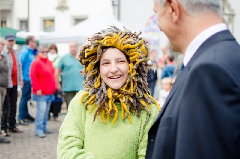 KI generiert: Das Bild zeigt eine lächelnde Frau mit einem auffälligen, löwenähnlichen Kopfschmuck aus gelben und braunen Dreadlocks, die an einem öffentlichen Ort mit anderen Menschen im Hintergrund steht. Der Hauptinhalt des Bildes ist die Frau mit dem ungewöhnlichen und farbenfrohen Accessoire.