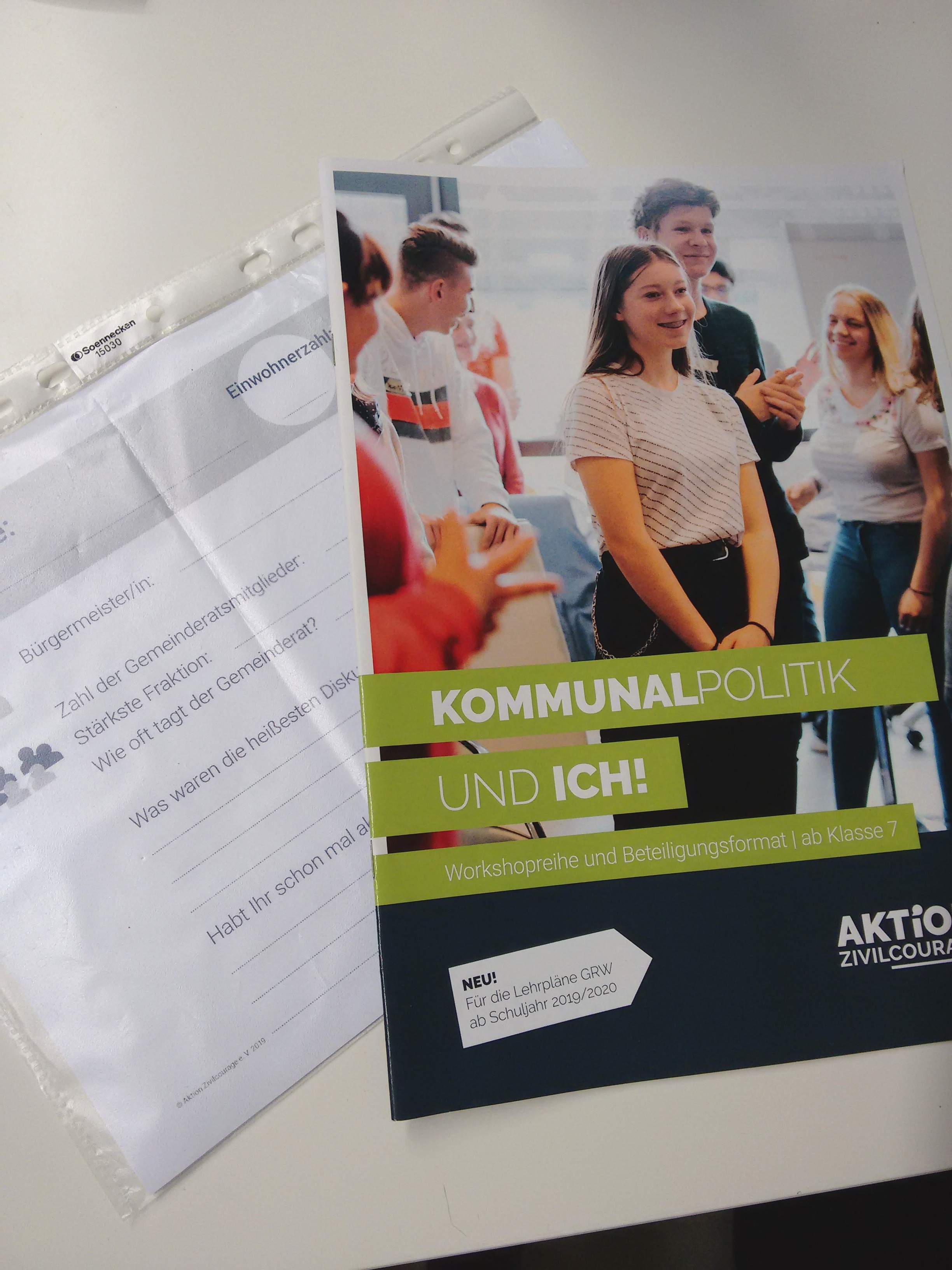 KI generiert: Das Bild zeigt ein Heft mit dem Titel "Kommunalpolitik und ich!" auf der Titelseite, das sich an Schüler ab Klasse 7 richtet und von der Aktion Zivilcourage herausgegeben wird. Neben dem Heft liegt ein leeres Blatt Papier mit einigen handschriftlichen Notizen und einem Stempel.