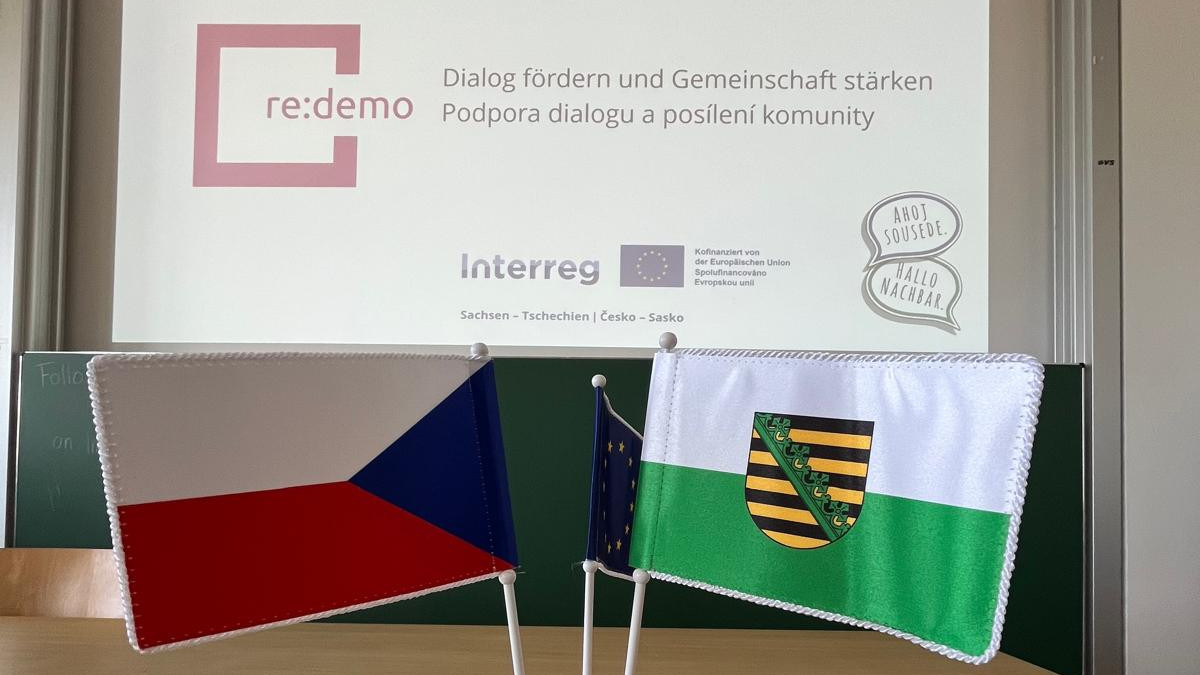 KI generiert: Das Bild zeigt eine Präsentation mit dem Titel "Dialog fördern und Gemeinschaft stärken" (Podpora dialogu a posílení komunity) und den Logos von re:demo und Interreg. Im Vordergrund sind zwei Flaggen zu sehen: die Flagge der Tschechischen Republik und die Flagge des Freistaates Sachsen.