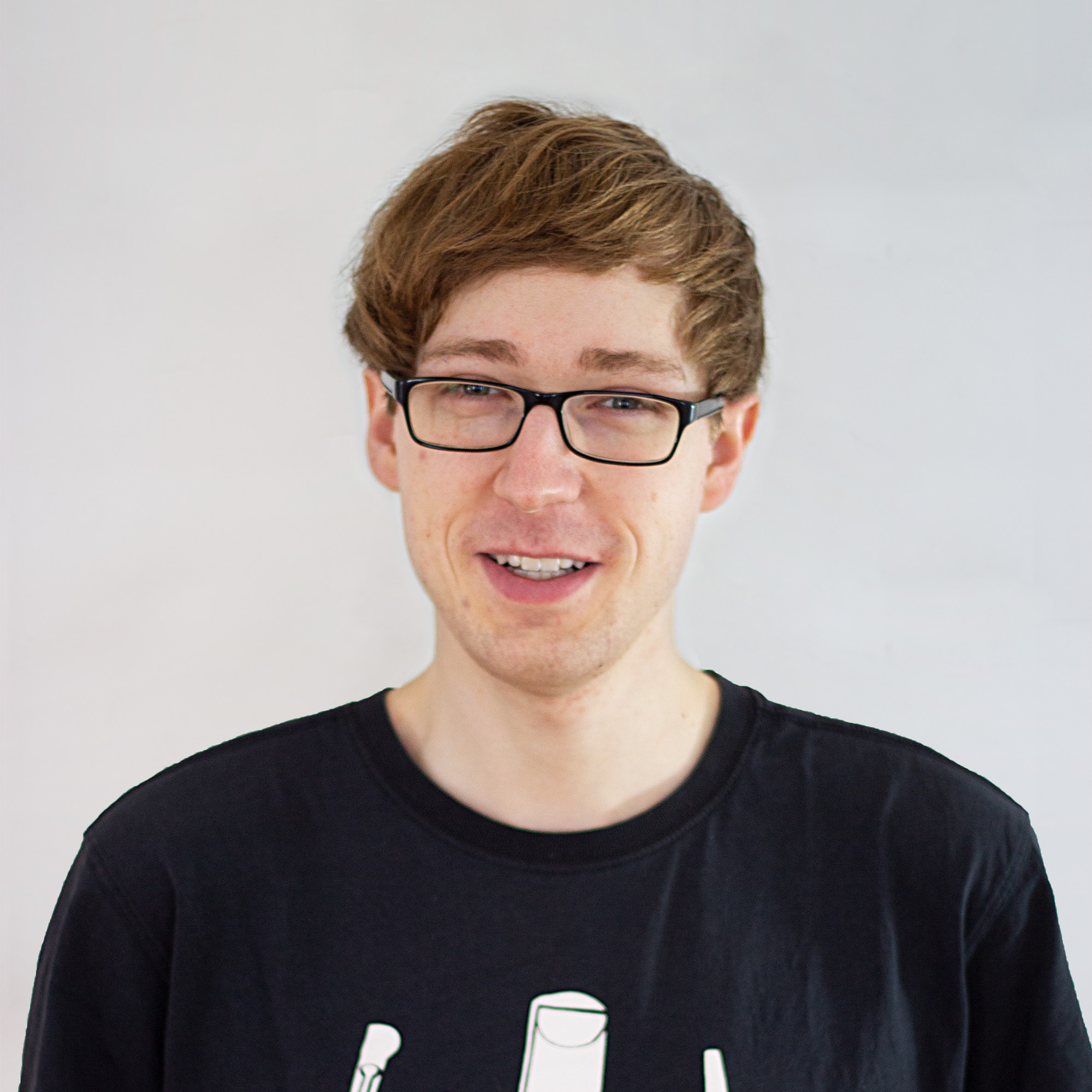 KI generiert: Das Bild zeigt einen lächelnden Mann mit kurzen, rötlichen Haaren und einer Brille, der ein schwarzes T-Shirt mit einem weißen Aufdruck trägt. Der Hauptinhalt des Bildes ist das Porträt des Mannes vor einem hellen, neutralen Hintergrund.