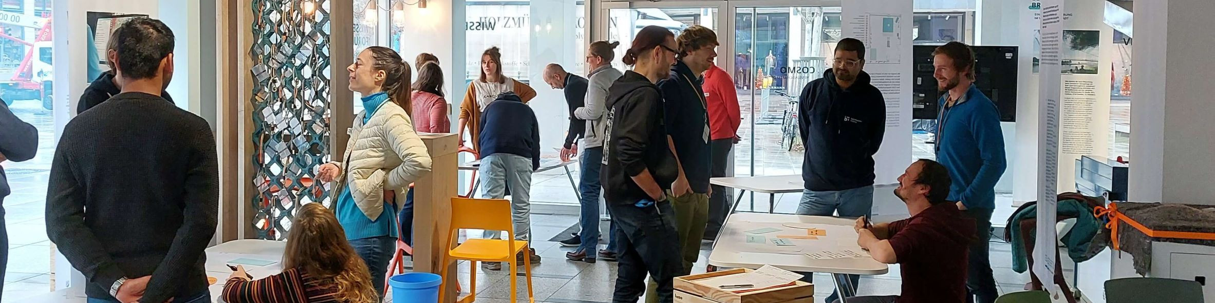KI generiert: Das Bild zeigt eine Gruppe von Menschen in einem hellen, modernen Innenraum, die sich unterhalten und an Tischen stehen. Der Hauptinhalt des Bildes ist eine soziale Zusammenkunft oder ein Event in einem Gemeinschaftsraum oder Büro.
