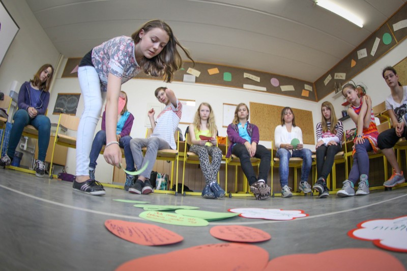 KI generiert: Das Bild zeigt eine Gruppe von Jugendlichen in einem Klassenzimmer, wobei eine Person im Vordergrund eine Aktivität auf dem Boden durchführt, bei der bunte Papierformen mit Aufschriften verteilt sind. Der Hauptinhalt des Bildes ist eine interaktive Gruppenaktivität oder ein Spiel, an dem die Jugendlichen teilnehmen.