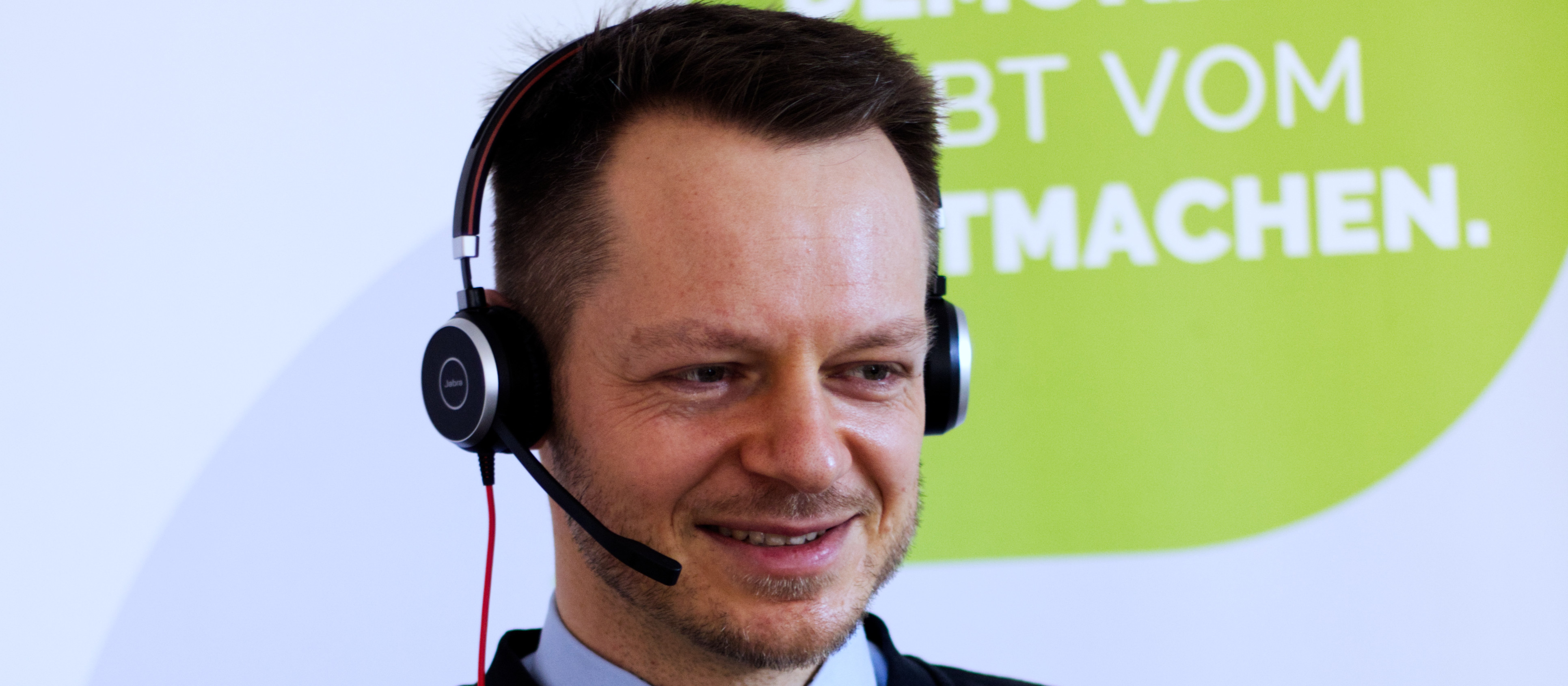 KI generiert: Das Bild zeigt einen lächelnden Mann mit Kopfhörer und Mikrofon vor einem Hintergrund mit teilweise lesbarer Aufschrift. Der Hauptinhalt des Bildes ist der Mann, der möglicherweise in einer Kundenservice- oder Callcenter-Umgebung arbeitet.
