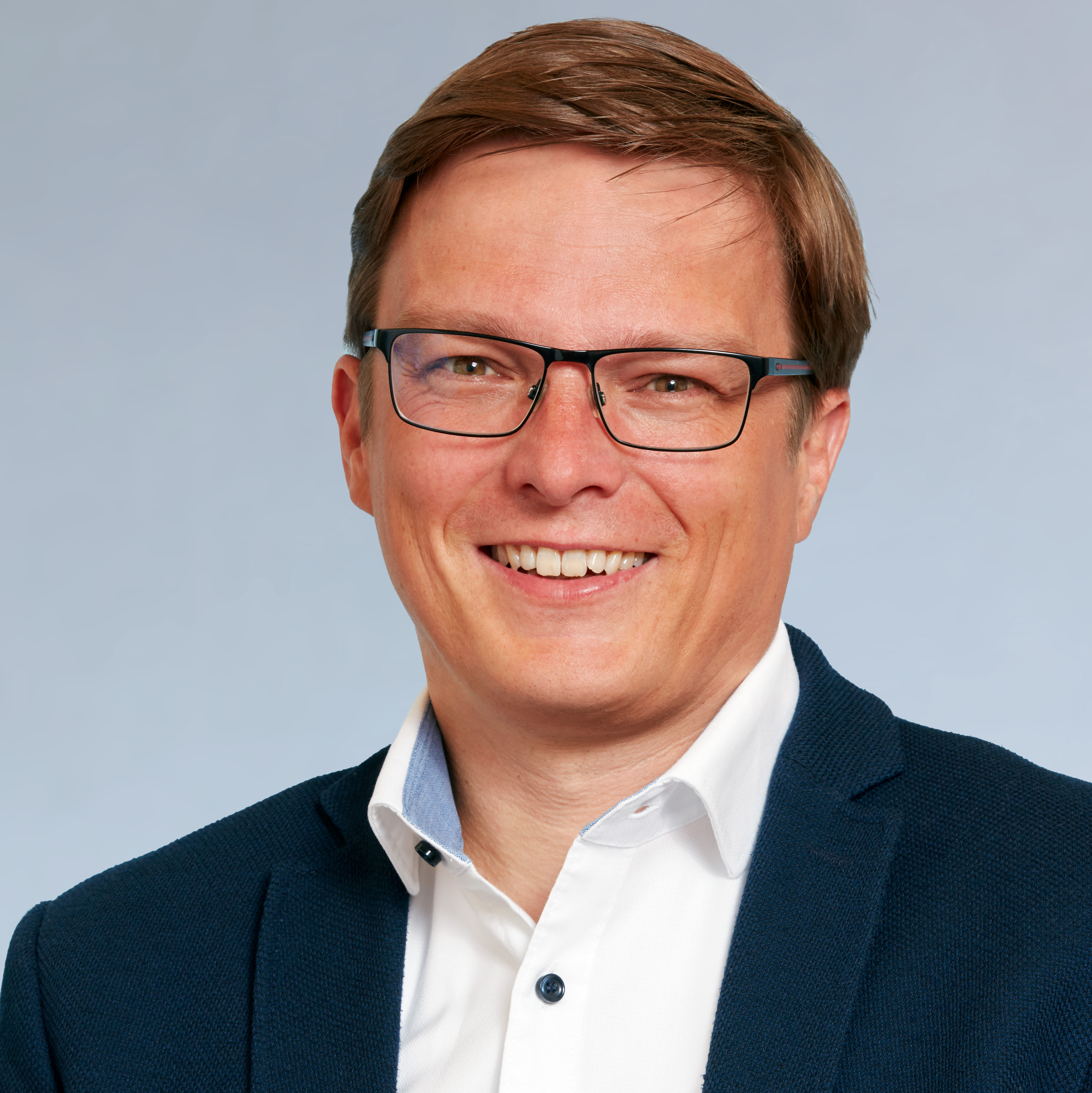 KI generiert: Das Bild zeigt einen lächelnden Mann mit kurzen braunen Haaren und einer Brille. Er trägt ein weißes Hemd mit einem dunklen Blazer und einem blauen Kragen.